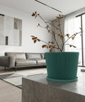 A teal planter pot with a plant sits on a dark wooden table next to a gray couch with a blanket.