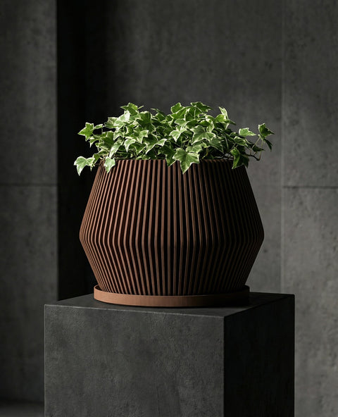 A Stromanthe plant in a brown, fluted planter pot sits atop a light wood cabinet. The plant has green and pink striped leaves. Next to the planter are three stacked books and another plant in a gray pot. A framed piece of art hangs on the wall behind the plants.