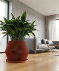 A close-up shot showcases a vibrant green plant nestled in a terracotta-colored planter pot. The pot features a unique vertical ribbed design, adding texture and visual interest. The plant and pot are placed on a wooden surface, with a large window providing natural light in the background. A minimalist shelf with decorative objects is visible in the corner, complementing the modern interior design.