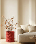 A red textured planter pot with a plant with brown leaves sits next to a beige couch and wooden table in a neutral room.