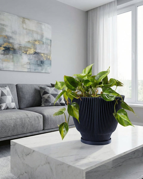 A black planter pot with a plant sitting on a marble coffee table in a living room. The living room has a gray couch with patterned pillows and a large painting on the wall. There is a window with white curtains in the background.