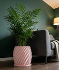 A pink planter pot with a green plant inside sits next to a gray couch in a room with dark green walls.