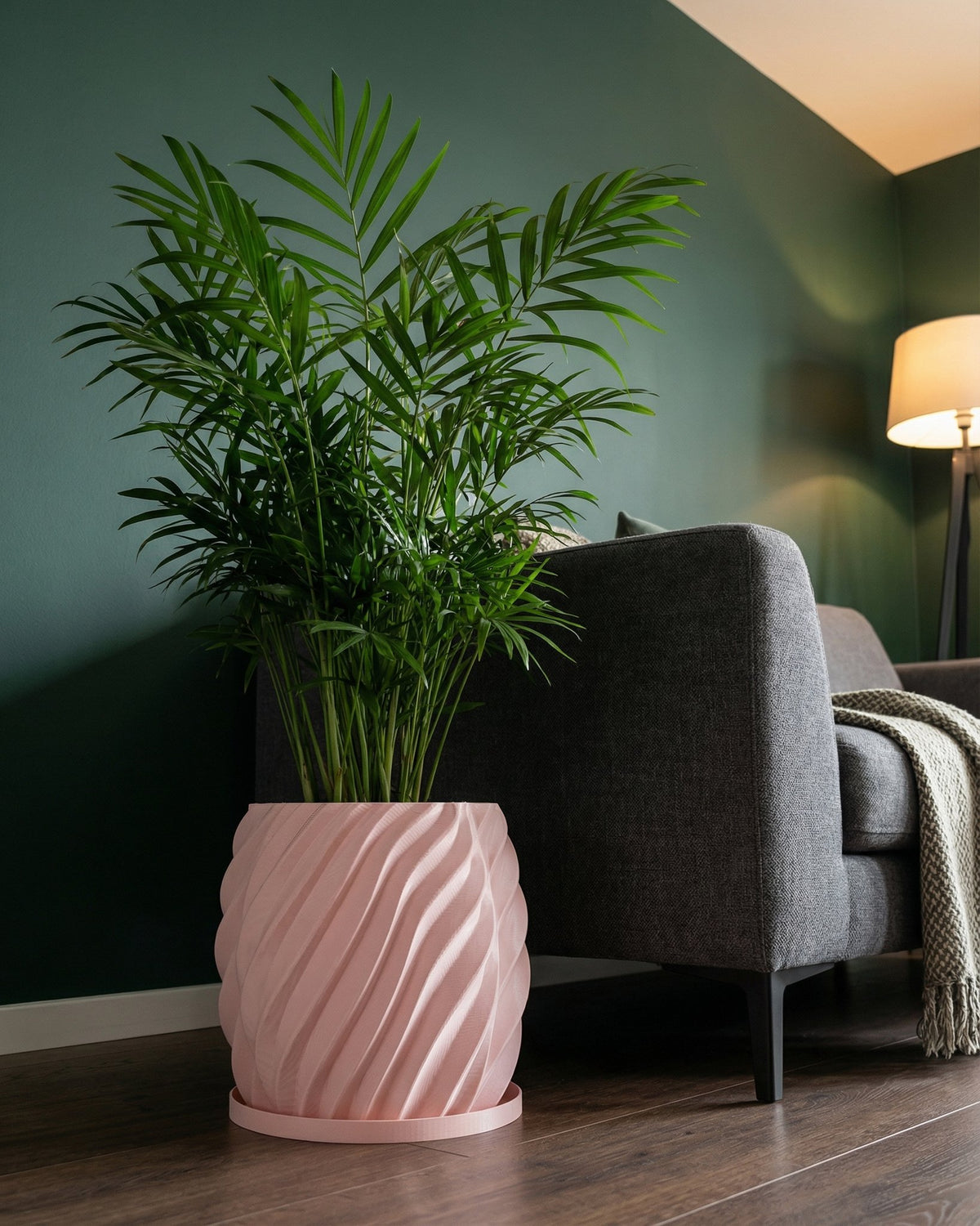 A pink planter pot with a green plant inside sits next to a gray couch in a room with dark green walls.