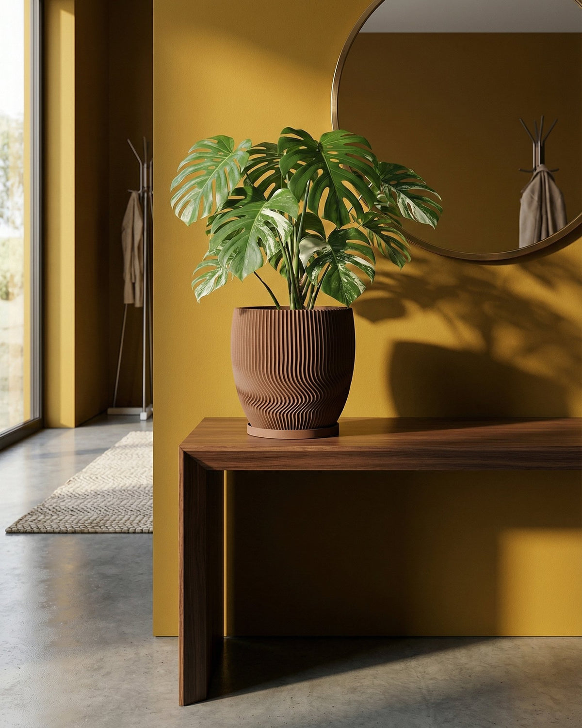 A Monstera plant sits in a brown, ridged planter pot on a wooden table against a yellow wall with a round mirror.