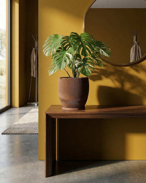 A Monstera plant sits in a brown, ridged planter pot on a wooden table against a yellow wall with a round mirror.