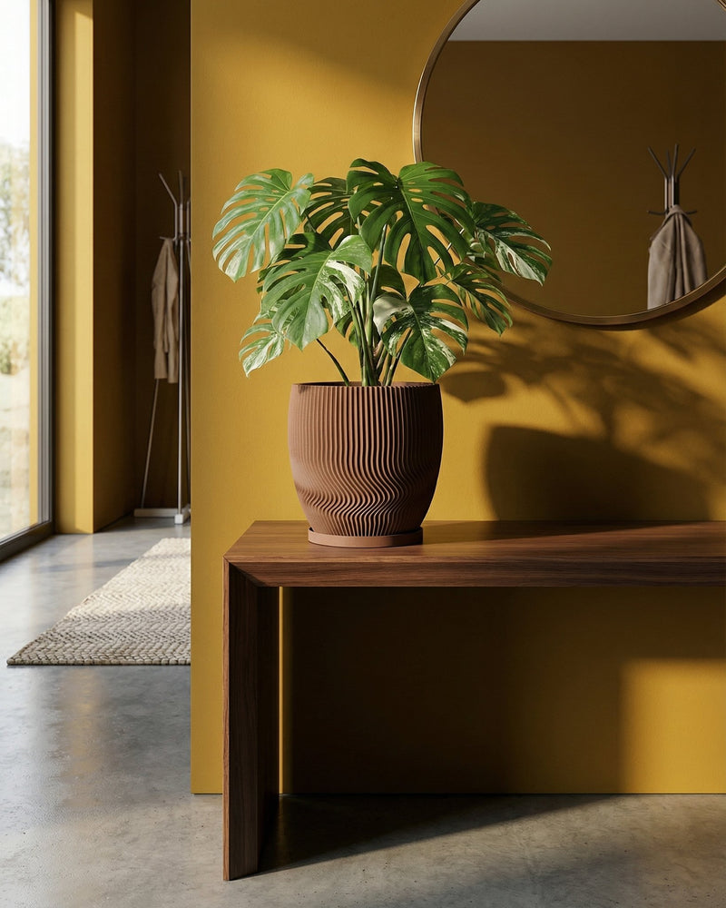 A Monstera plant sits in a brown, ridged planter pot on a wooden table against a yellow wall with a round mirror.