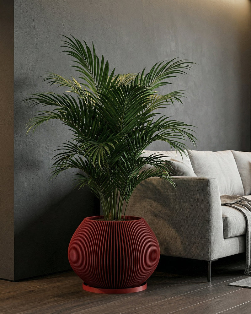 A tall, vibrant green Monstera plant is potted in a striking, vertically ribbed red planter. The planter sits atop a modern, light-wood table with a unique, open base design. The setting is a bright, minimalist living space with a gray sofa, abstract art, and sheer white curtains.