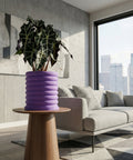 A purple planter pot with a plant on a wooden table in a living room with a couch and a city view.