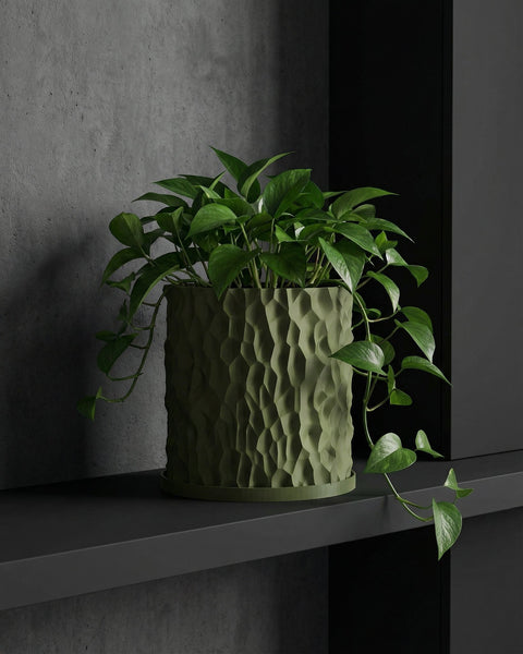 A green pothos plant in a textured green planter pot sits on a dark gray shelf against a gray wall.