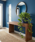 A blue planter pot with a green plant sits on a wooden console table in front of a blue wall with a round mirror.