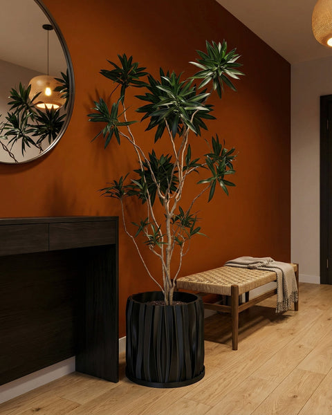 A black planter pot with a plant sits on a dark wood table. A round mirror hangs on the wall behind the plant.