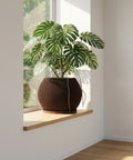 A Monstera plant in a brown, ribbed planter pot sits on a wooden windowsill, bathed in natural light. The pot is the focal point.