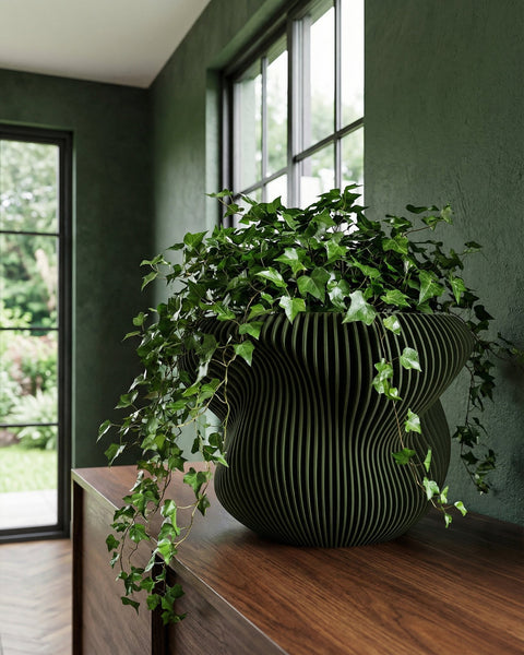 A green planter pot with a plant sits on a wooden shelf in front of a window. The pot has a ribbed design.