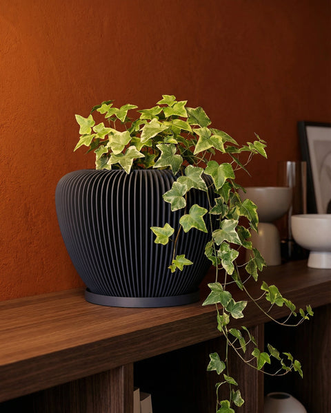 A black planter pot with a plant on a wooden shelf. The pot has a unique ribbed design.
