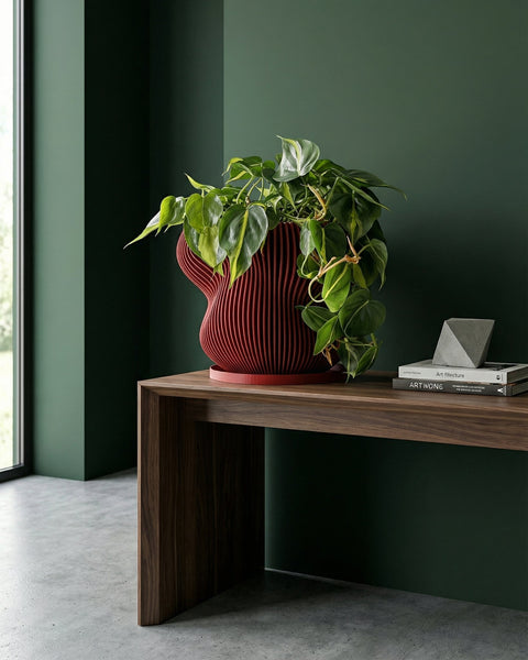 A red planter pot with a plant in it sits on a wooden surface next to a window, casting a shadow on the wall.