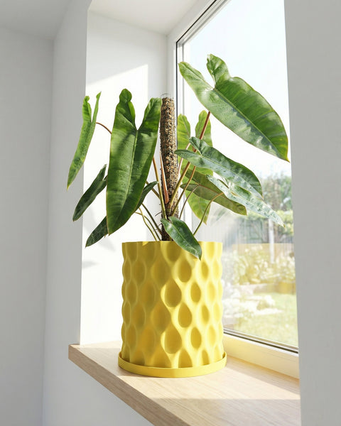 A yellow planter pot with a plant inside sits on a windowsill in front of a window.