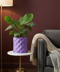 A purple planter pot with a green plant sits on a marble table next to a gray couch with a blanket.