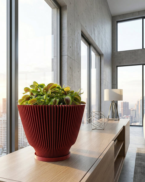 A red planter pot with a Venus flytrap plant inside sits on a light wood console table in front of a large window with a city view. The pot has a ribbed design and a flared base. A geometric metal sculpture and a white lamp are also on the table. The room has a modern, minimalist aesthetic with concrete walls and large windows.