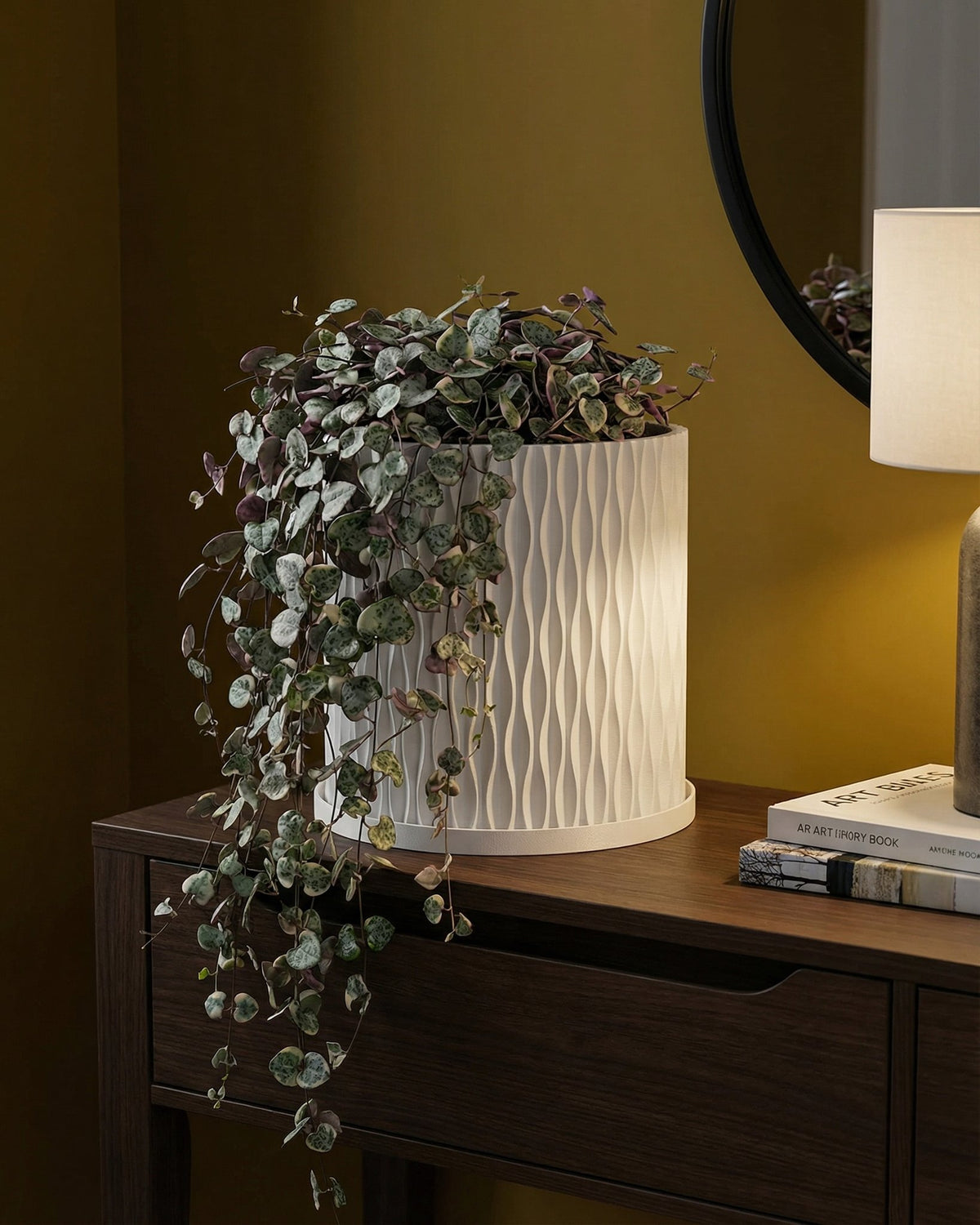 A string of pearls plant spills from a white, textured planter pot on a wooden console table in a room with dark green walls.