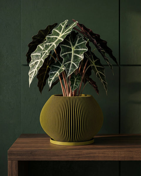 A green planter pot with a plant is placed on a wooden platform in a bedroom. The bed is in the background with gray bedding and a yellow blanket. A modern lamp sits on a nightstand next to the bed. A large window is on the left side of the image.