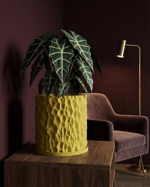 A yellow planter with a green plant sits on a wooden table. A chair and lamp are in the background.