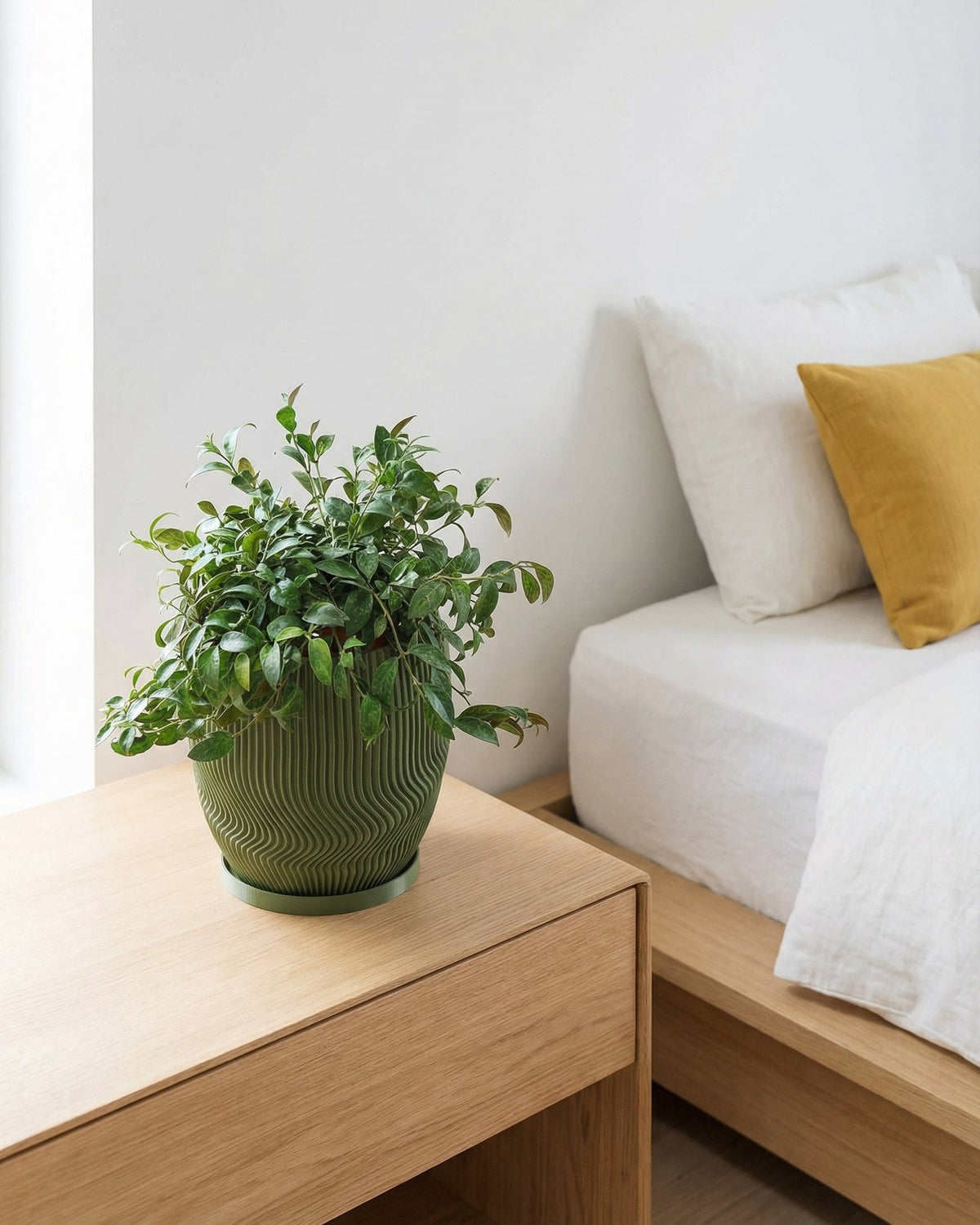 A green planter pot with a plant sits on a wooden nightstand next to a bed with white linens and a yellow pillow.