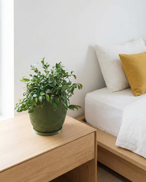 A green planter pot with a plant sits on a wooden nightstand next to a bed with white linens and a yellow pillow.