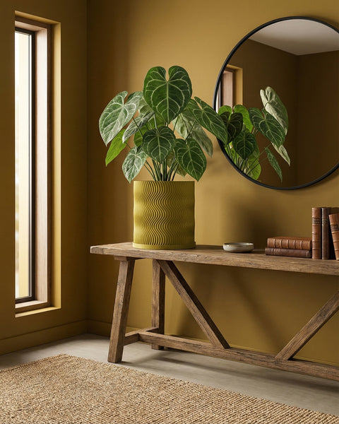 A plant in a wavy green planter pot sits on a wooden table next to a round mirror and books in a room with mustard-colored walls.