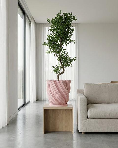 A pink planter pot with a green plant sits on a wooden table next to books and a gold lamp.