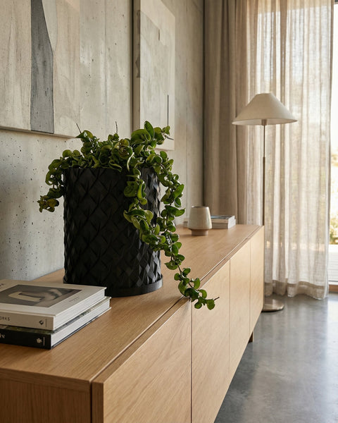 A pothos plant in a black planter sits on a wooden windowsill, bathed in sunlight. The planter has a woven texture.