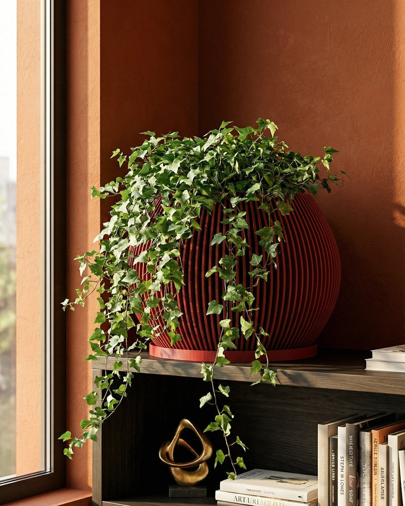A red, round, slatted planter pot filled with a lush green ivy plant sits on a dark wood shelf next to books and a gold sculpture.