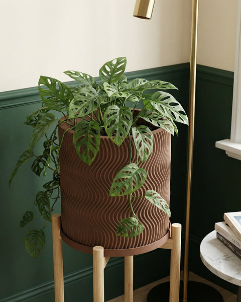 A brown planter pot with a wavy design sits on a wooden stand, holding a lush green plant with unique holey leaves.