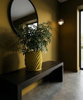 A green plant in a yellow, swirled planter pot sits on a dark wood table against a yellow wall with a round mirror.