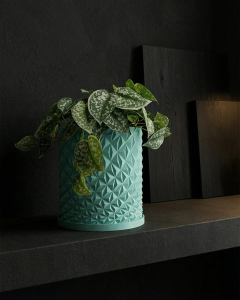 A teal geometric planter pot with a green plant sits on a dark shelf against a dark wall.