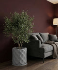 A gray planter pot with a diamond pattern sits next to a gray couch in a room with dark wood floors and a burgundy wall.