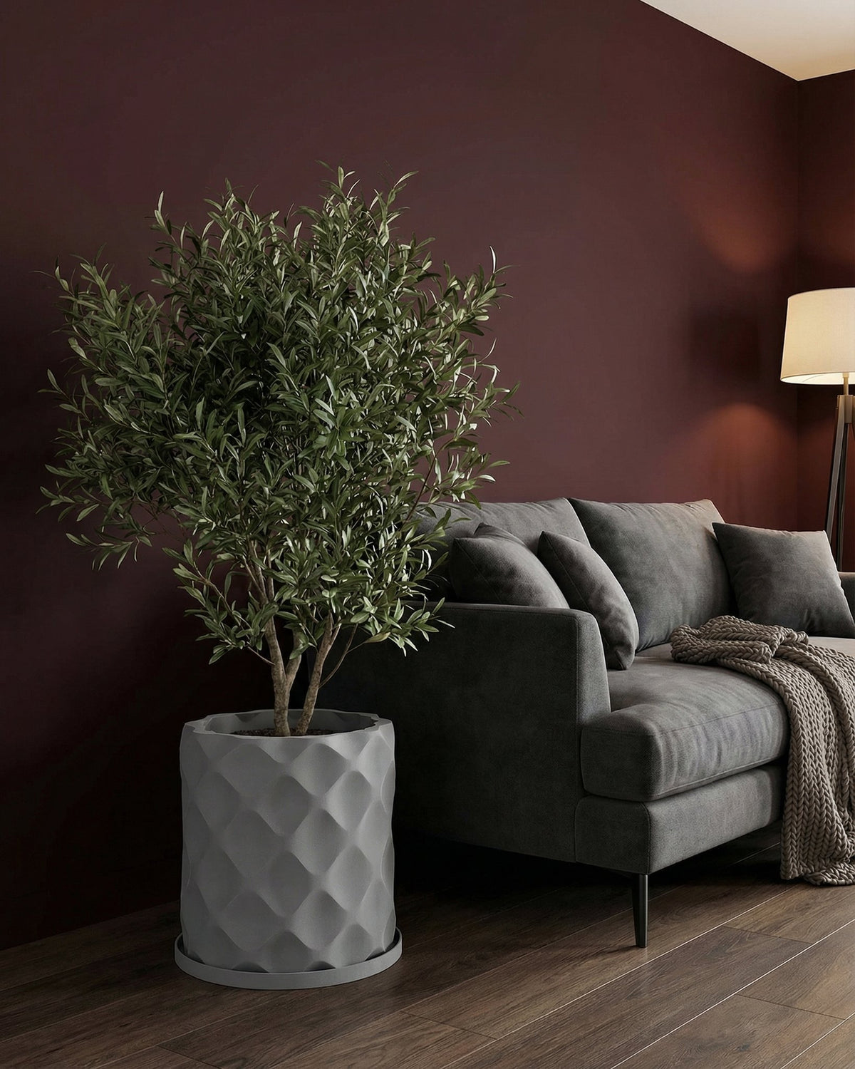 A gray planter pot with a diamond pattern sits next to a gray couch in a room with dark wood floors and a burgundy wall.