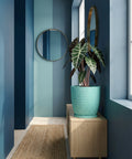 A turquoise planter pot with a plant sits on a wooden cabinet in a blue-themed room with a round mirror.