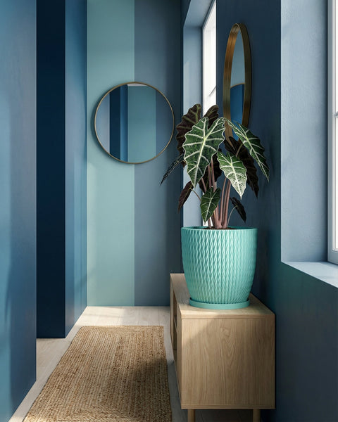 A turquoise planter pot with a plant sits on a wooden cabinet in a blue-themed room with a round mirror.