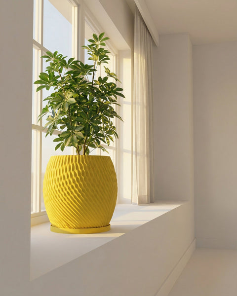 A bright yellow planter pot with a green plant sits on a white windowsill next to a window with sheer white curtains.