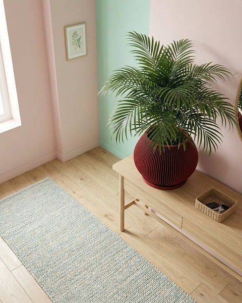 A red planter pot with a Venus flytrap plant inside sits on a light wood console table in front of a large window with a city view. The pot has a ribbed design and a flared base. A geometric metal sculpture and a white lamp are also on the table. The room has a modern, minimalist aesthetic with concrete walls and large windows.