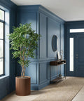 A brown, ribbed planter pot with a green tree inside sits in a blue entryway with a rug and console table.