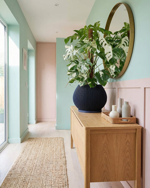 A black planter pot with a plant sitting on a marble coffee table in a living room. The living room has a gray couch with patterned pillows and a large painting on the wall. There is a window with white curtains in the background.