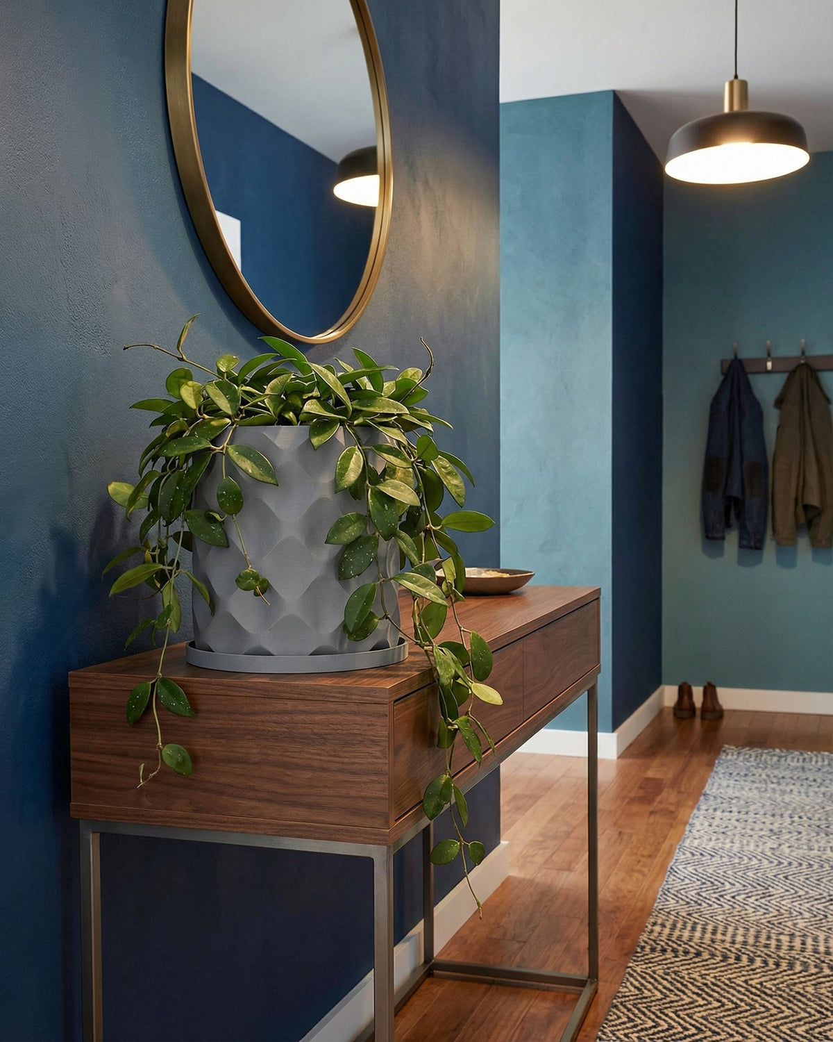 A gray planter pot with a diamond pattern sits atop a wooden console table in a stylish entryway. The pot is filled with a lush green plant.