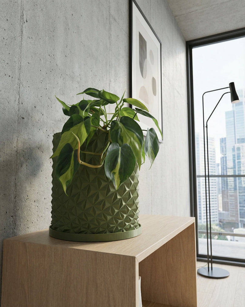 A green planter pot with a geometric design sits on a light wood shelf, with a plant inside. A framed print and a window with a city view are in the background.
