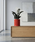A red planter pot with a geometric pattern sits on a wooden cabinet in a modern room with a painting on the wall.