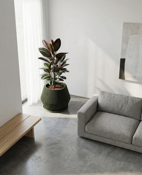 A green Anthurium plant in a green planter pot sits on a light-colored wooden table in a modern living room. The planter has a unique, vertically-lined design. A gray wall, a framed geometric print, and a chair are in the background.