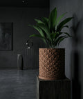 A brown geometric planter pot with a green plant sits on a wooden block in a dark room.