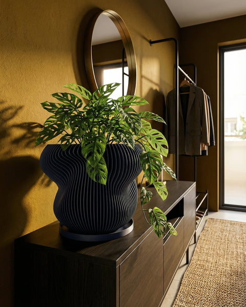 A black planter pot with vertical lines holds a small tree with green leaves in a dimly lit room.