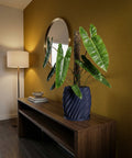 A blue planter pot with a plant inside sits on a wooden table in a room with a mirror and a lamp.