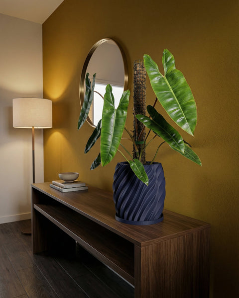 A blue planter pot with a plant inside sits on a wooden table in a room with a mirror and a lamp.